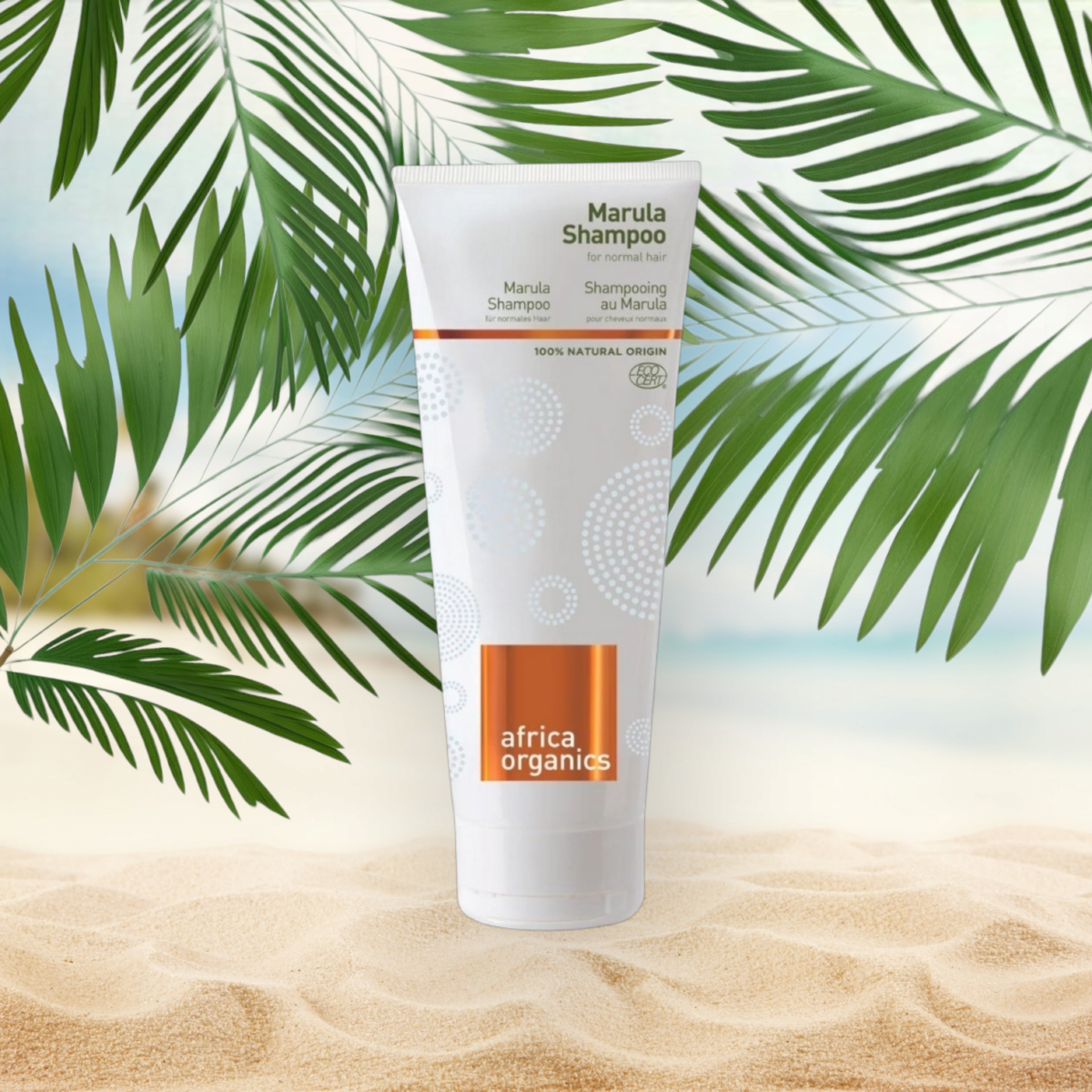 Pureday En tube Africa Organics Marula shampoo 210ml står på en sandstrand baggrund, delvist indrammet af grønne palmeblade, under en klar himmel. Røret er hvidt med orange accenter og tekst, der beskriver dets funktioner.