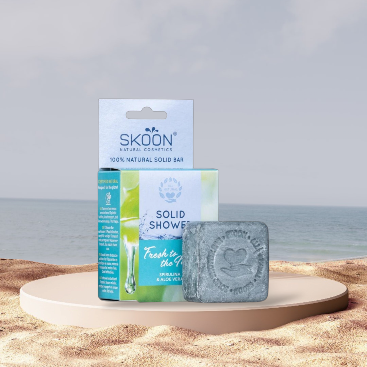 Pureday En Skoon solid brusestang og dens emballage vises på en beige bakke med sandstrand og hav i baggrunden. Sæbe baren har et hjerteaftryk og en grå farve, der understreger dens naturlige ingredienser.